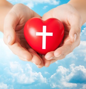 religion, christianity and charity concept - close up of female hands holding red heart with christian cross symbol over blue sky and clouds background